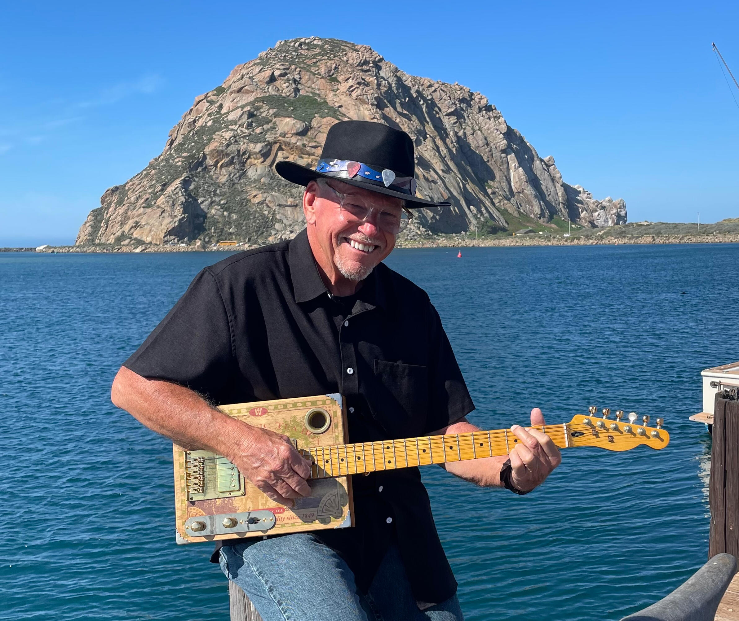 CigarBoxRay Photograph of Ray Riordan playing one of his cigar box guitars in front of a brown fence with a market umbrella, sailboat masts, bay, and a sandbar behind the fence. At Ray's feet are two additional cigar box guitars on stands and a speaker on a stand to h